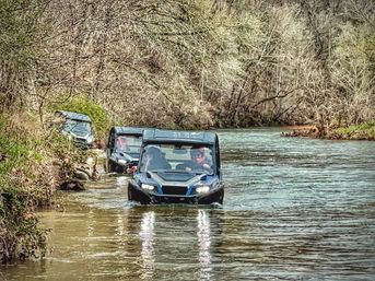 Two off-road UTVs splashing through a shallow river on a wooded trail, headlights reflecting on the water and bare trees lining the banks — outdoor off-road adventure.