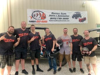 Eight men in matching “Wolf Pack” jerseys smiling and holding helmets, posing by off-road side-by-side vehicles on a gravel lot in front of a metal building with a rentals banner.