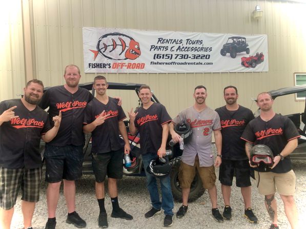 Eight men in matching “Wolf Pack” jerseys smiling and holding helmets, posing by off-road side-by-side vehicles on a gravel lot in front of a metal building with a rentals banner.