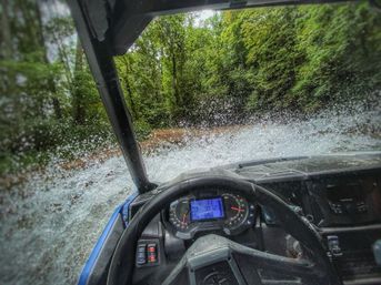 Cockpit view of an off-road UTV splashing through a shallow forest stream, water spraying over the windshield and hood, dashboard and steering wheel visible with lush green trees along the trail.