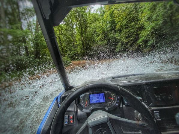 Cockpit view of an off-road UTV splashing through a shallow forest stream, water spraying over the windshield and hood, dashboard and steering wheel visible with lush green trees along the trail.