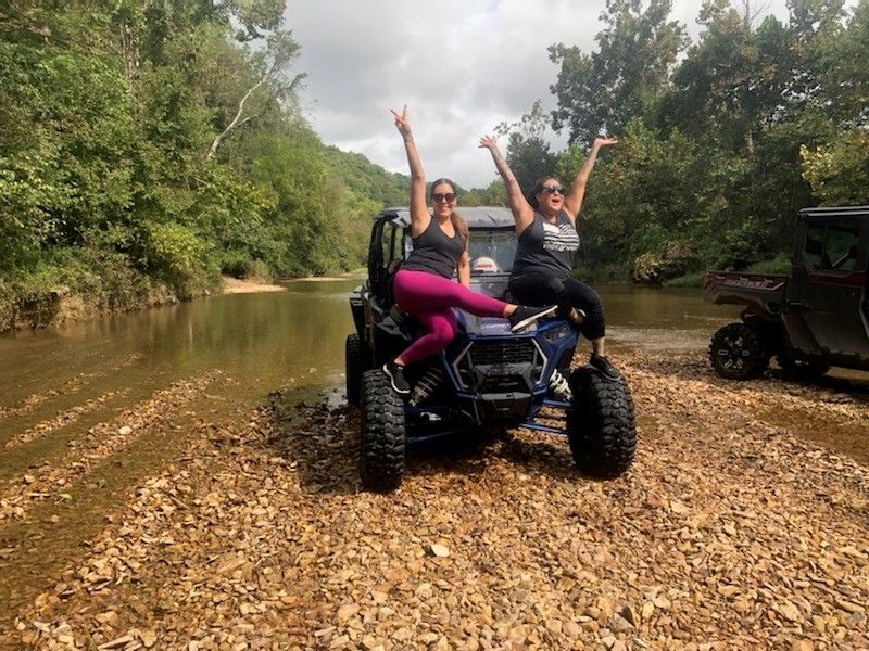 Two friends cheering atop a blue off-road UTV parked on a rocky riverbank beside a shallow creek, surrounded by green forest — outdoor adventure.