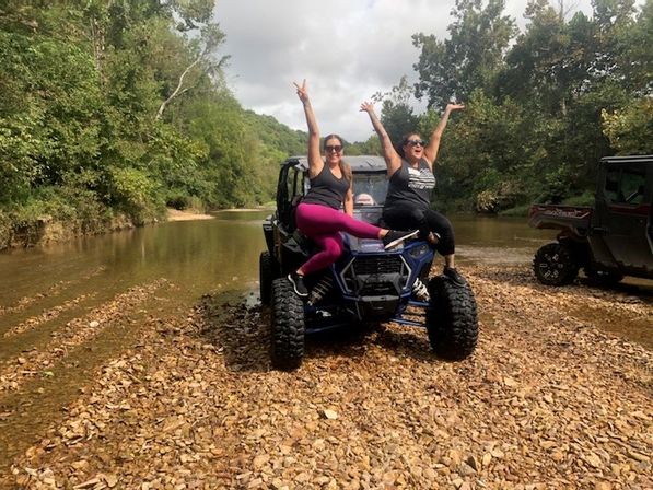 Two friends cheering atop a blue off-road UTV parked on a rocky riverbank beside a shallow creek, surrounded by green forest — outdoor adventure.