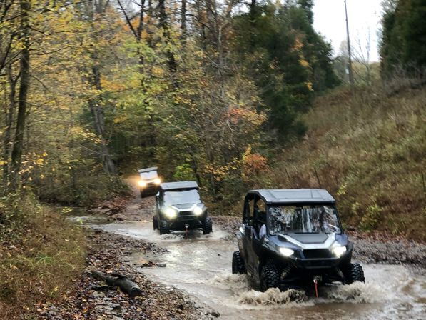 Three side-by-side off-road UTVs splashing through a shallow muddy stream on a narrow wooded trail surrounded by autumn foliage.