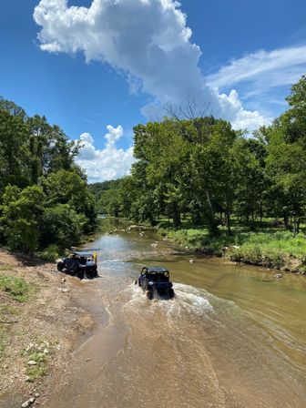 Two off-road UTVs splashing through a shallow creek on a wooded trail, surrounded by lush green trees under a bright blue sky with billowing white clouds