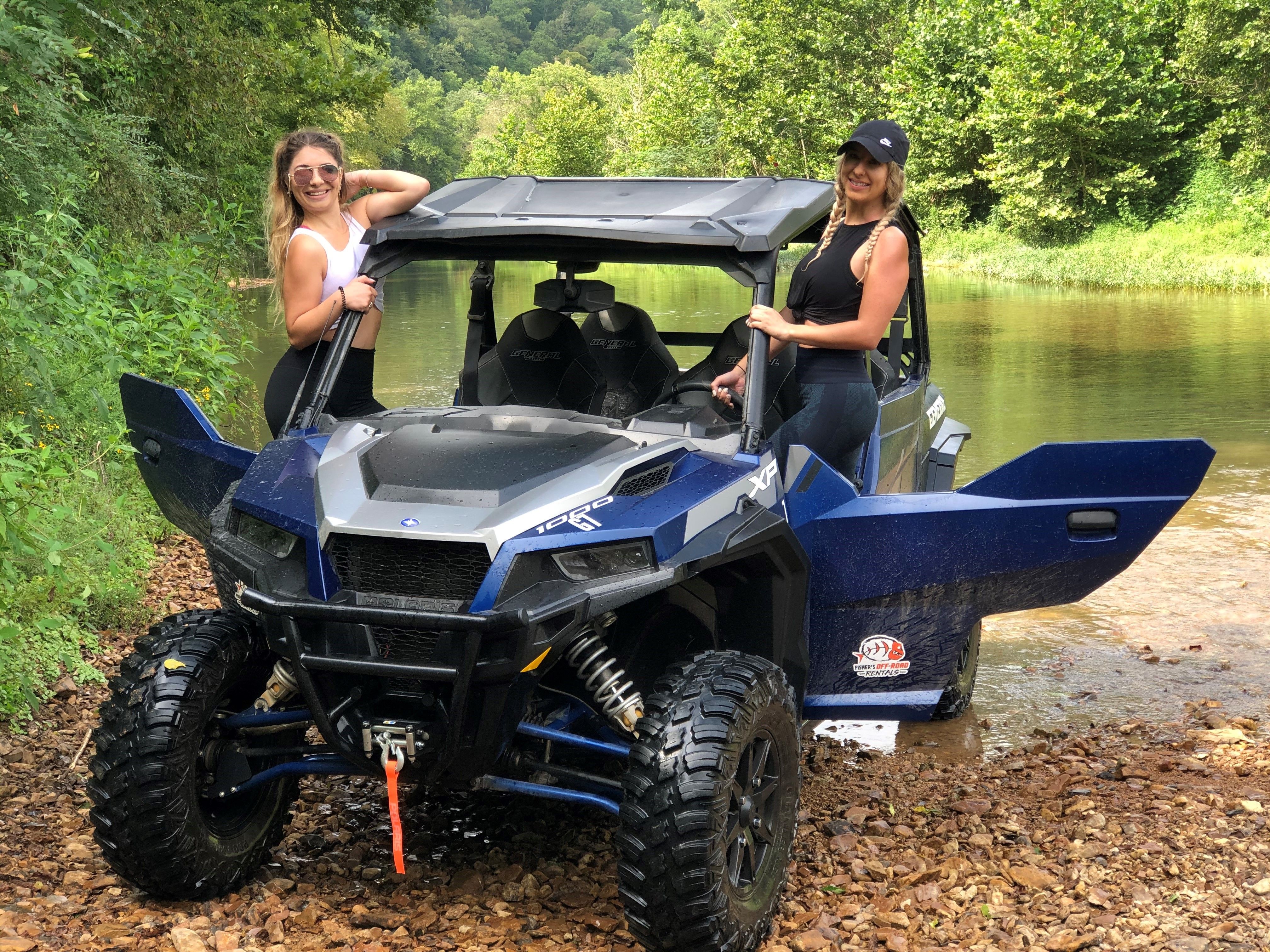 Two women smiling and posing with a blue off-road side-by-side UTV parked on a rocky creek bank, calm river and green forested hills in the background — outdoor off-road adventure.
