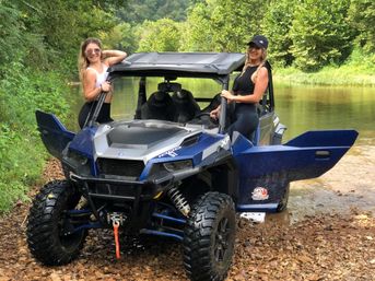 Two women smiling and posing with a blue off-road side-by-side UTV parked on a rocky creek bank, calm river and green forested hills in the background — outdoor off-road adventure.