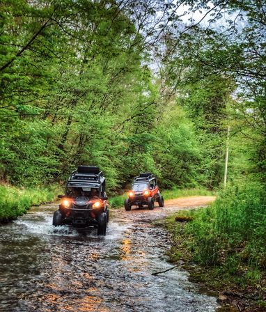 Two off-road UTVs splashing through a shallow forest stream on a muddy dirt trail, headlights glowing, surrounded by lush green trees and spring foliage.