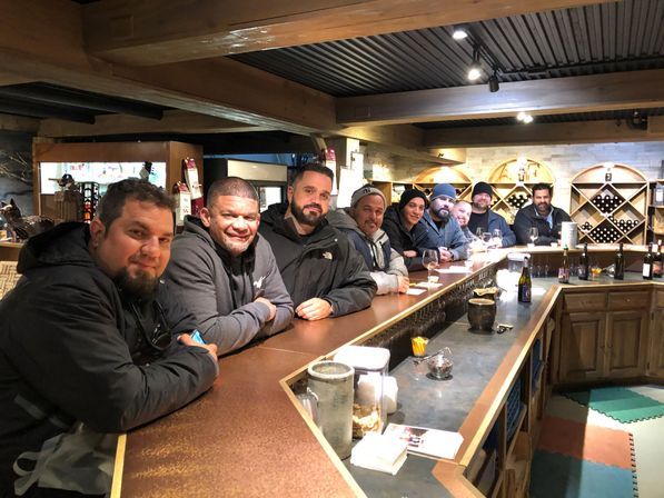 Group of men lined up along a rustic wooden wine-tasting bar with wine glasses, bottles, and cozy cabin-style interior