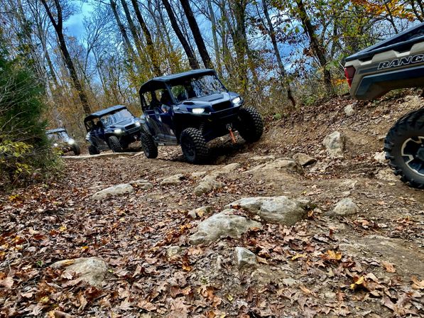 UTV convoy tackling a rocky, leaf-covered forest trail in autumn — off-road side-by-side vehicles on a wooded dirt path