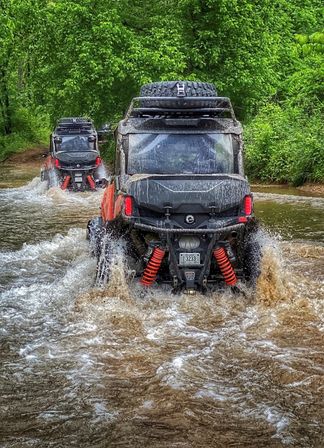 Two off-road UTVs fording a muddy forest creek, orange suspension springs and roof spare tire visible as water splashes around tires on a green woodland trail — outdoor off-road adventure.