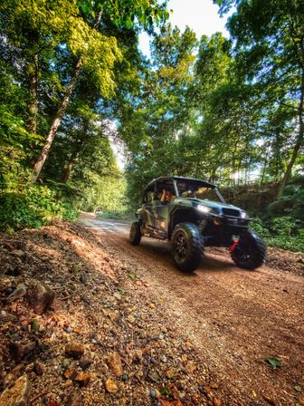 Side-by-side UTV on a rocky forest trail, off-road adventure on a sunlit, wooded gravel road with trees overhead and kicked-up dirt.