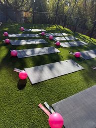 Sunlit outdoor fitness terrace with artificial turf, rows of gray yoga mats each paired with a pink mini exercise ball and resistance bands on a fenced deck surrounded by trees.