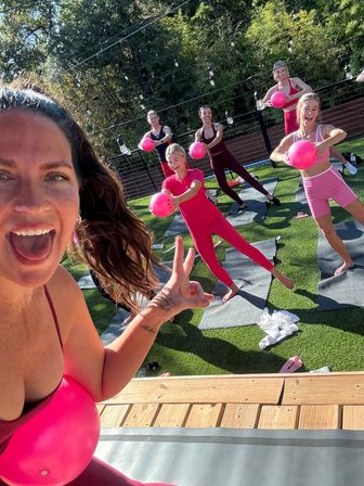 Fun outdoor group fitness class on a sunny lawn with women in pink activewear holding bright pink exercise balls on yoga mats under string lights
