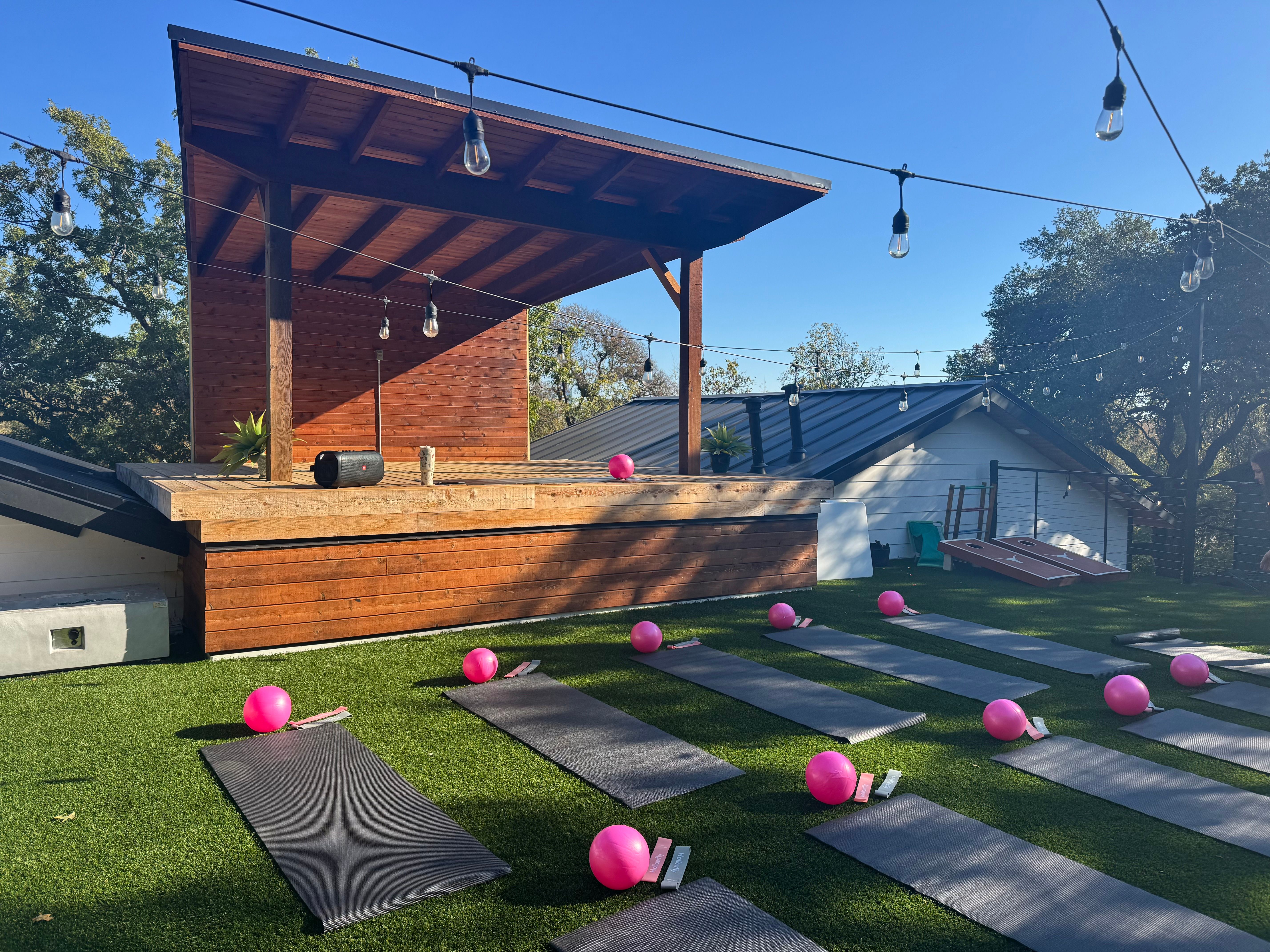 Sunlit rooftop fitness deck with a wooden covered stage and rows of black yoga mats on artificial turf, each paired with a bright pink exercise ball beneath hanging string lights and a clear blue sky.
