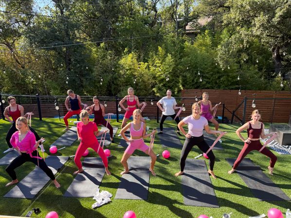 Energetic outdoor group fitness class on a sunny lawn — women in pink and purple activewear using resistance bands and pink exercise balls on yoga mats beneath string lights and trees.