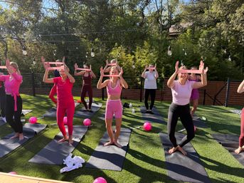 Sunny outdoor group fitness class on artificial turf: women in pink workout clothes on yoga mats holding pink rectangular props up to their faces like binoculars, with pink exercise balls, string lights and trees in the background.
