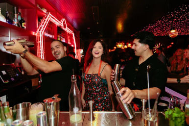 Two bartenders shake cocktail shakers while a smiling woman stands between them at a neon-lit downtown bar, surrounded by bottles, glassware and festive string lights in a lively nightlife scene.