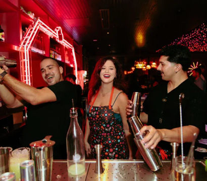Two bartenders shake cocktail shakers while a smiling woman stands between them at a neon-lit downtown bar, surrounded by bottles, glassware and festive string lights in a lively nightlife scene.