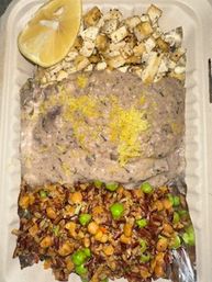 Takeout container with a lemon wedge and diced grilled cubes, a large scoop of creamy dip topped with lemon zest, and a seasoned grain-and-vegetable medley with edamame and corn.