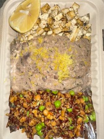 Takeout container with a lemon wedge and diced grilled cubes, a large scoop of creamy dip topped with lemon zest, and a seasoned grain-and-vegetable medley with edamame and corn.