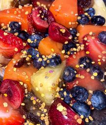 Close-up of vibrant fruit bowl with blueberries, strawberries, halved cherries, pineapple and orange melon chunks sprinkled with bee pollen