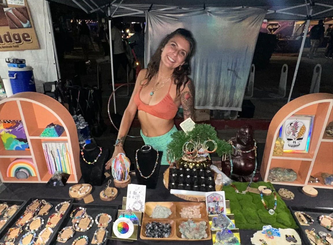 Smiling vendor at an outdoor night market booth displaying handmade beaded necklaces, bracelets, earrings and crystals on colorful shelves and trays.