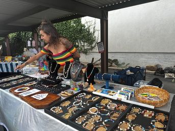 Artisan jewelry booth at an outdoor market — colorful handmade beaded bracelets and necklaces on trays and display busts, vendor in a rainbow off-shoulder top arranging pieces under a covered pavilion.