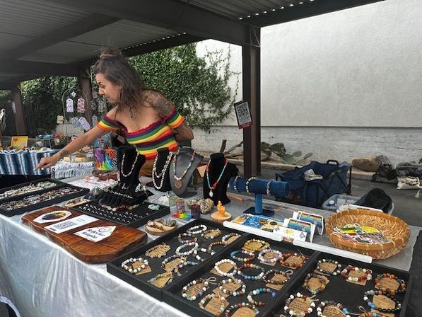 Artisan jewelry booth at an outdoor market — colorful handmade beaded bracelets and necklaces on trays and display busts, vendor in a rainbow off-shoulder top arranging pieces under a covered pavilion.
