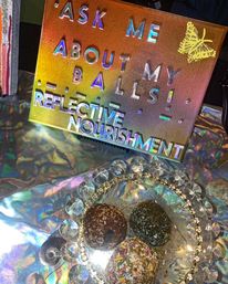 Holographic rainbow sign reading Ask me about my balls — Reflective Nourishment above a crystal-rimmed plate with three coconut- and sprinkle-coated energy balls on a shimmering metallic tablecloth.