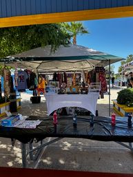 Outdoor craft market booth under a canopy tent on a sunny day with a palm tree, clothing racks, a white display table of handmade goods, and a picnic table in the foreground with paint bottles and craft supplies.