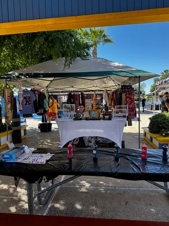 Outdoor craft market booth under a canopy tent on a sunny day with a palm tree, clothing racks, a white display table of handmade goods, and a picnic table in the foreground with paint bottles and craft supplies.
