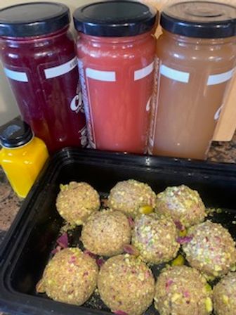 Tray of pistachio‑coated energy balls on a kitchen counter beside three tall cold-pressed juice bottles (deep red, pink, amber) and a small yellow juice bottle — healthy snack and juice combo.