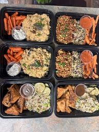Six black meal-prep containers on a countertop with portions of seasoned rice, chopped cabbage slaw, roasted baby carrots, golden-brown chicken pieces, and small dipping sauce cups — colorful healthy prepared lunches.