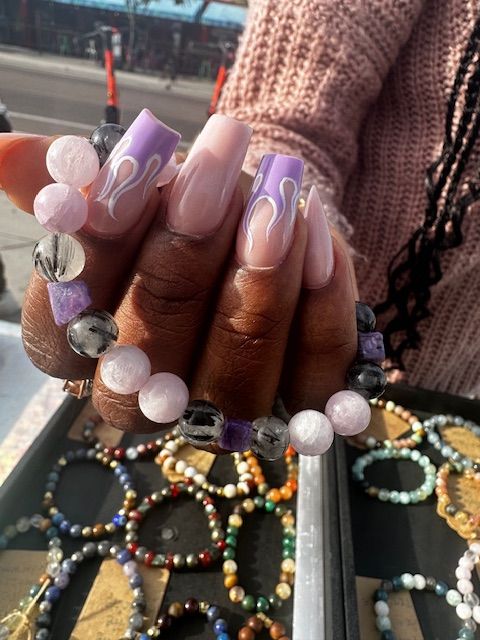 Hand with long nude-to-lavender coffin acrylic nails with white flame tips holding pink and gray beaded gemstone bracelets over a tray of colorful beaded bracelets at an outdoor market stall