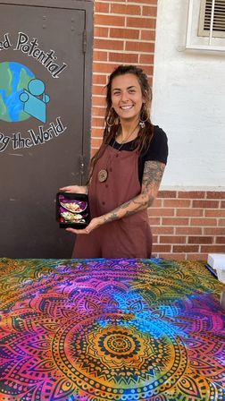 Smiling food vendor in a brown apron holding a black container of purple cabbage tacos at an outdoor market booth with a brick wall and a vibrant rainbow mandala tablecloth.