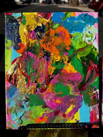 Close-up of a vibrant abstract impasto canvas painting with thick textured strokes of orange, pink, green, blue and metallic gold, propped on an outdoor easel.