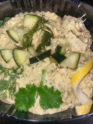 Creamy Mediterranean-style hummus topped with cucumber cubes, pickled dill, cilantro leaves and a lemon wedge in a plastic takeout container