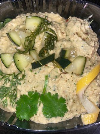 Creamy Mediterranean-style hummus topped with cucumber cubes, pickled dill, cilantro leaves and a lemon wedge in a plastic takeout container