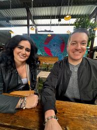 Two smiling friends at a wooden table on a covered outdoor patio with a large colorful bird mural, hanging pendant lights and casual dining vibe.