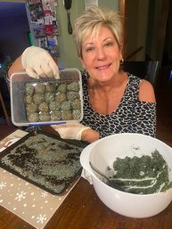 Smiling woman at a kitchen table holding a plastic container of hand-rolled green herb-coated balls, wearing gloves with a white mixing bowl of green mixture and a tray with herb crumbs nearby.