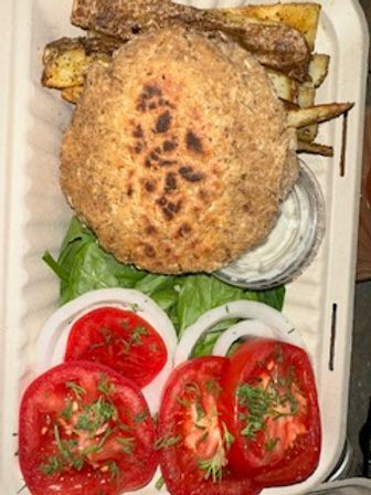 Takeout container with a toasted burger bun resting on golden fries, crisp lettuce, sliced tomatoes and onion rings, and a small cup of creamy dipping sauce