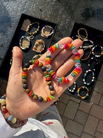 Sunlit hand holding two handmade colorful beaded bracelets — rainbow chip beads and natural stone rounds — over a display tray of more bracelets at an outdoor market.