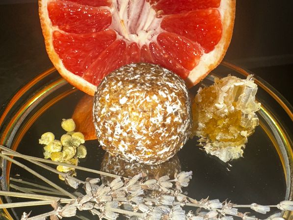 Halved pink grapefruit, coconut-coated energy ball, honeycomb chunk, chamomile buds and dried lavender arranged on a reflective black plate — bright citrus and herbal snack.