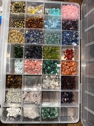 Clear plastic compartment organizer filled with colorful gemstone and crystal beads — polished chips, pearls and round beads neatly sorted for jewelry making and craft projects in a craft studio.