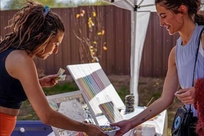 Two women exchanging cash and a small item at an outdoor artisan market stall with colorful prints and handmade goods displayed on a table under a pop-up canopy.
