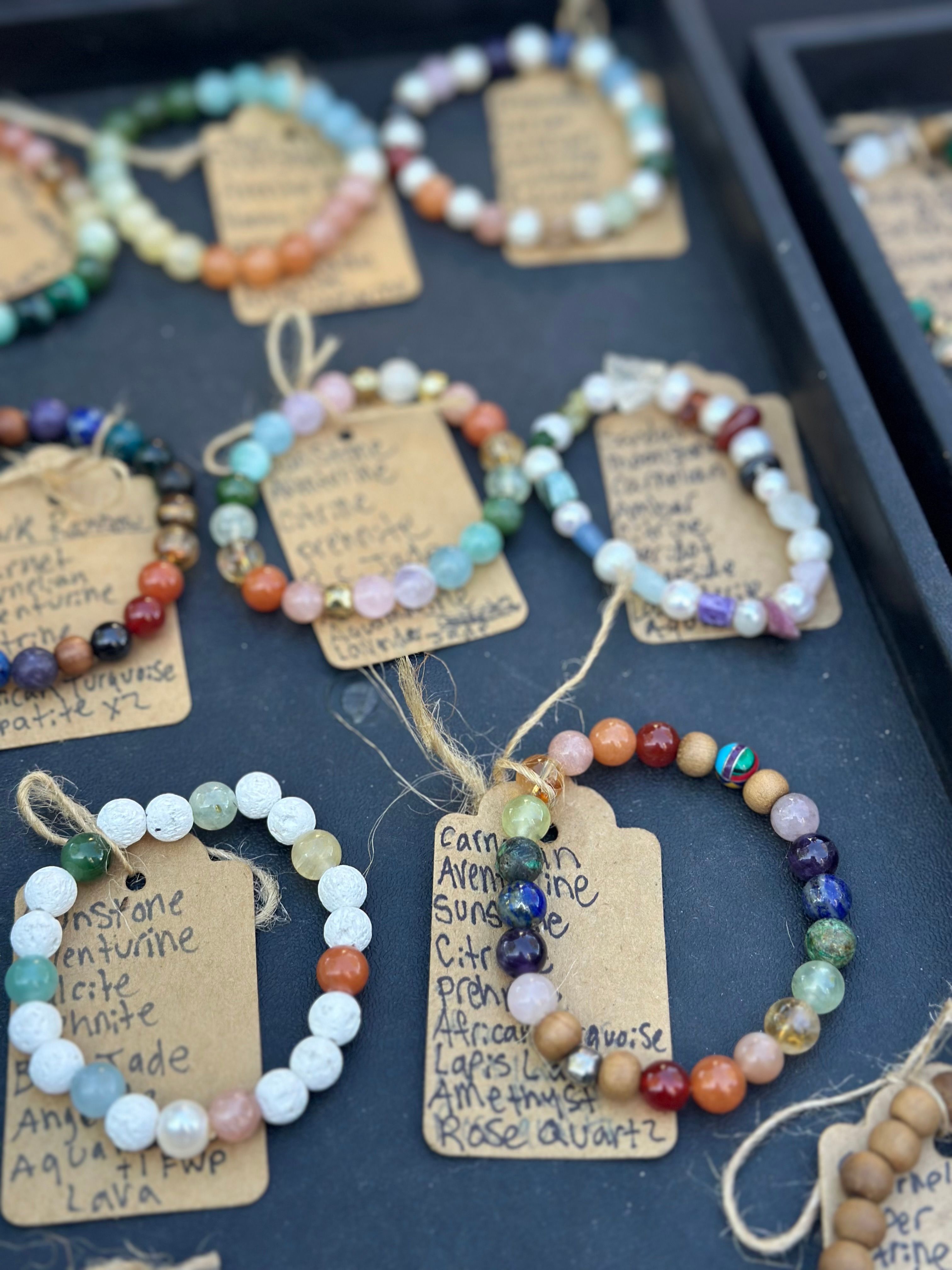 Colorful handmade gemstone and lava bead bracelets on kraft paper tags arranged on a black display tray at an artisan market stall