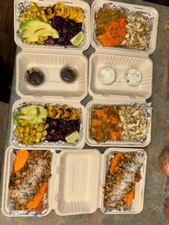 Colorful meal-prep to-go boxes on a counter with healthy bowls of roasted sweet potato and shredded chicken, grilled plantains, avocado slices, black beans, corn, purple cabbage, lime wedges and small sauce cups — ready-to-eat takeout lunches.