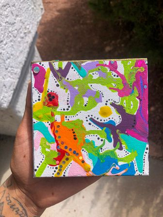 Hand holding a small square canvas painted with vibrant neon abstract acrylic splashes and dotted black accents on a white background, photographed outdoors in sunlight.