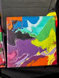 Colorful abstract acrylic pour canvas with eye-catching swirls of purple, teal, lime, yellow, orange and black, displayed outdoors on a small stand.